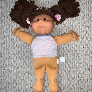 Puede incluir: Muñeca Cabbage Patch Kids con cabello de hilo marrón peinado en dos coletas altas. La muñeca tiene ojos marrones, una expresión sonriente y lleva una camiseta sin mangas blanca con un estampado floral rosa y ribete azul.