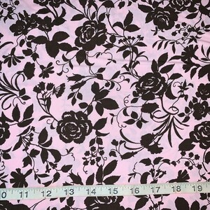 May include: Pink fabric with a repeating pattern of dark brown roses and foliage. The floral design covers the entire surface. A ruler is placed across the bottom of the fabric, showing measurements in inches.