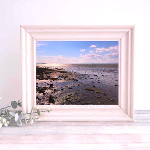 May include: A framed photograph of a beach scene with a sailboat in the distance. The beach is sandy and wet, with the tide going out. The sky is blue with white clouds. The frame is white.