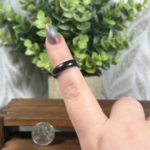 May include: A black, glossy ring is worn on a finger. The ring is a solid band with a smooth surface. A silver coin is visible in the foreground. Green foliage is in the background.
