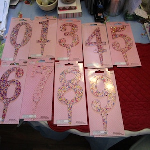 May include: Set of ten pink cake toppers with numbers 1 through 9 and 0. Each number is decorated with colorful sprinkles and has a small stick for inserting into a cake.