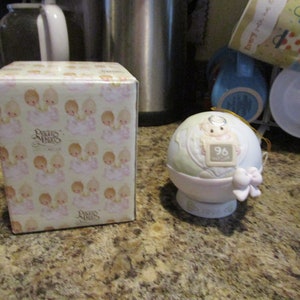 May include: A white porcelain Precious Moments ornament shaped like a globe with a cherub angel holding a sign that says "96 Years". The ornament is on a white base and has a gold ribbon. The ornament is in a box with a pattern of cherub angels.