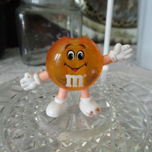 May include: Orange M&M character figurine with a smiling face and the M&M logo on its chest. The figurine has white arms and legs and is standing on a clear glass surface.