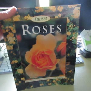 May include: A book titled "Roses" by Sunset. The cover features a large, orange rose with a dark background. The border is decorated with yellow flowers and green leaves. The book is held by a person.