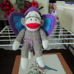May include: A sock monkey plush toy featuring a grey and white knit body, a bright red mouth, and a purple hat. The monkey has large purple and blue felt wings attached to its back and a thin purple string tied around its waist as a belt.
