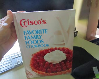 May include: A cookbook titled "Crisco's Favorite Family Foods Cookbook" is held in a hand. The cover features a pie with a red filling and a dollop of whipped cream. The title is in blue and red lettering.