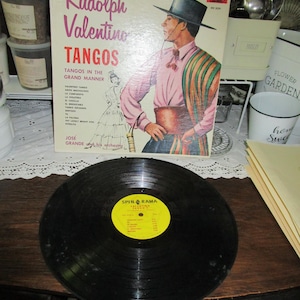 May include: A vintage vinyl record album cover featuring Rudolph Valentino with the title "Tangos" in bold red letters. The album cover is a vibrant pink, green, and yellow color scheme. The album features a photo of Rudolph Valentino in a black hat and a striped shirt. The album cover also includes a list of tango songs.