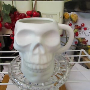 May include: A matte white ceramic mug shaped like a human skull. The mug features a hollow interior, a curved handle on the right side, and a detailed molded surface depicting eye sockets, a nasal cavity, and teeth. The skull mug sits on a clear glass base.