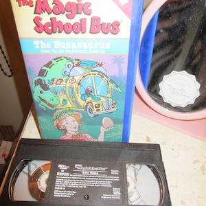 May include: A VHS tape of the Magic School Bus episode "Gets Eaten" with a blue and yellow bus on the cover. The cover also features a girl holding a dinosaur egg. The title "The Busasaurus" is printed on the cover.