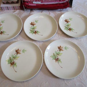 May include: Five off-white ceramic plates with a leaf design in shades of brown and green. The plates are arranged on a white tablecloth. The leaf design is on the left side of each plate.