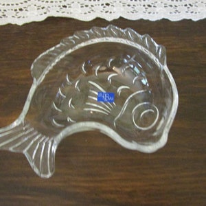 May include: A clear glass fish-shaped dish with a raised design. The dish has a blue sticker on the side that says "CRYSTAL."