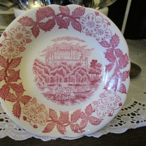 May include: A white ceramic saucer with a pink floral design around the edge. The center features a pink illustration of a castle surrounded by trees and a boat with people in it.