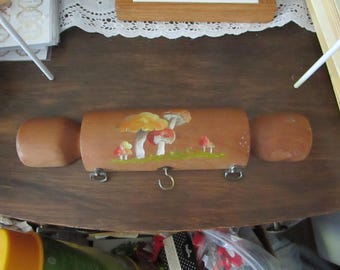 Könnte beinhalten: Ein Schlüsselhalter aus Holz in Form eines Baumstamms mit einem bemalten Design von Pilzen. Der Halter hat zwei Metallhaken an jeder Seite und einen einzelnen Haken in der Mitte.