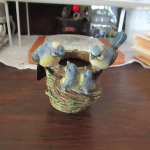 May include: A bluebird figurine perched on a nest with three baby bluebirds. The nest is made of twigs and leaves and is sitting on a wooden surface.