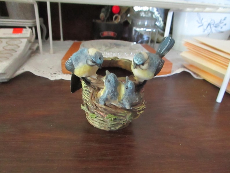 May include: A bluebird figurine perched on a nest with three baby bluebirds. The nest is made of twigs and leaves and is sitting on a wooden surface.