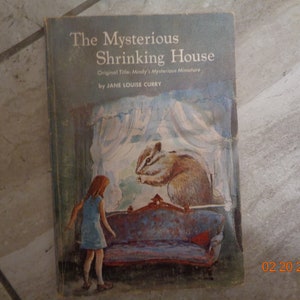 May include: A vintage children's book cover with a blue background. The title is "The Mysterious Shrinking House" with the original title "Mindy's Mysterious Miniature" listed below. The author is Jane Louise Curry. The cover illustration shows a girl in a blue dress looking at a squirrel sitting on a couch in a window.