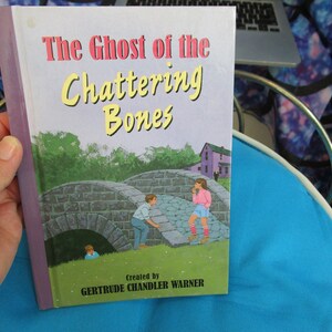 May include: A purple and white paperback book titled "The Ghost of the Chattering Bones" by Gertrude Chandler Warner. The cover features a stone bridge with two children standing on it.
