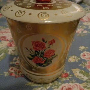 May include: A vintage gold and white metal cookie jar with a red lid and a floral design featuring pink roses. The jar has a swirl pattern around the sides.