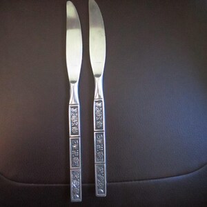 May include: Two silver-toned stainless steel dinner knives with serrated blades. The handles feature a rectangular design with three distinct panels, each embossed with a detailed floral pattern. The metal has a polished finish throughout.