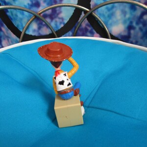 Pode incluir: Uma pequena figura de a&ccedil;&atilde;o do Woody da franquia Toy Story. A figura est&aacute; usando um chap&eacute;u de cowboy marrom, uma camisa branca com um padr&atilde;o de vaca preta, uma camisa amarela, cal&ccedil;as azuis e botas marrons. A figura est&aacute; sentada em um bloco bege.