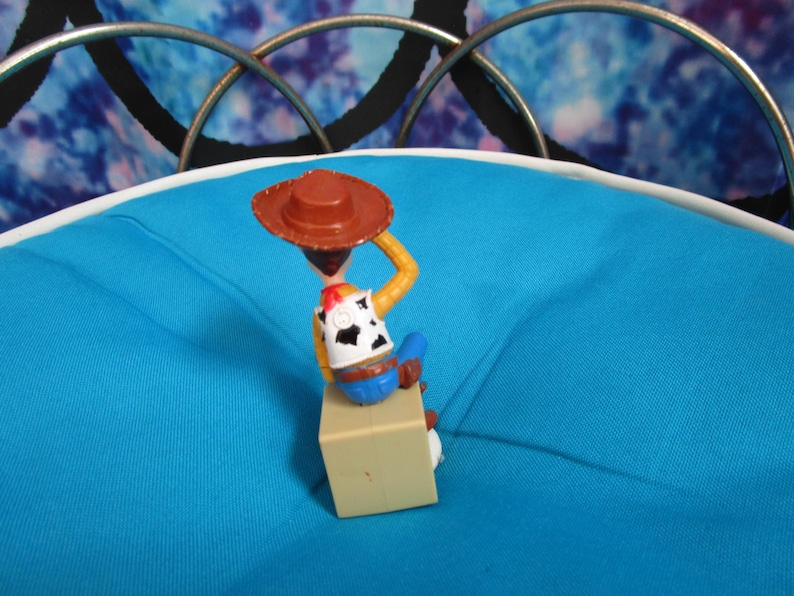 Pode incluir: Uma pequena figura de a&ccedil;&atilde;o do Woody da franquia Toy Story. A figura est&aacute; usando um chap&eacute;u de cowboy marrom, uma camisa branca com um padr&atilde;o de vaca preta, uma camisa amarela, cal&ccedil;as azuis e botas marrons. A figura est&aacute; sentada em um bloco bege.
