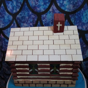 May include: A small wooden model of a log cabin with a white brick roof and a red cross on top. The cabin has three windows with stained glass designs.