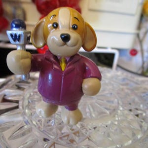 May include: A figurine of a cartoon dog wearing a purple suit and holding a microphone with the letter "W". The dog has tan ears and a tan and white head. The figurine is standing on a clear, patterned surface.