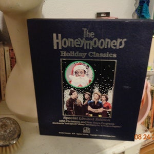 May include: A blue DVD case with a silver title, "The Honeymooners Holiday Classics: Special Limited Edition 1963 Christmas and New Years Programs". The cover features a photo of a man dressed as Santa Claus and a group of people standing in front of a snowy background.