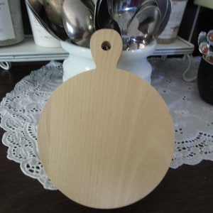 May include: A round wooden cutting board with a handle. The board is light brown and has a smooth surface.