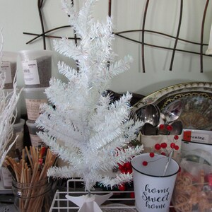 May include: A small white artificial Christmas tree with a white metal stand. The tree is decorated with iridescent glitter. A white metal bucket with black lettering that reads "home sweet home" is in the foreground.