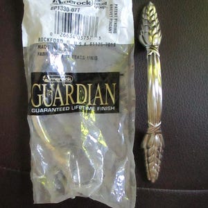 May include: A silver-toned metal cabinet pull with a textured wheat-like design at both ends. The handle is displayed next to its original clear plastic packaging, which features the Amerock Guardian brand name and product code BP1330-077.