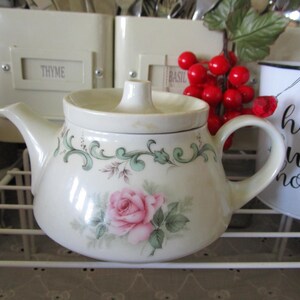 May include: A white teapot with a floral design featuring green leaves and pink roses. The teapot has a lid and a handle. The teapot is sitting on a white metal rack.