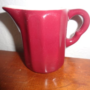 May include: A small, burgundy-colored ceramic creamer with a white interior and a white rim. The creamer has a rounded body and a handle.