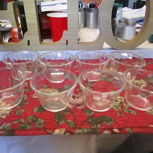 May include: Eight clear glass dessert bowls with a scalloped rim are arranged on a red floral patterned quilted placemat. The bowls are small and appear to be made of pressed glass. The background includes a wooden sign and other kitchen items.