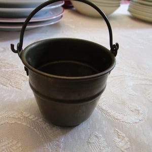 May include: A small, antique-style metal bucket with a dark bronze finish. It has a rounded body with two horizontal bands and a curved handle. The bucket is likely used for decorative purposes.