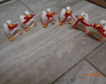 May include: Six white ceramic goose napkin rings with red bows. The geese are facing forward with their heads tilted slightly down. The bows are tied around the necks of the geese.