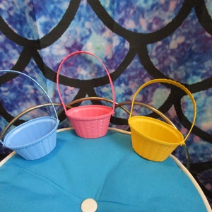 May include: Three small plastic baskets with woven designs. The baskets are blue, pink, and yellow. The baskets are sitting on a blue surface.