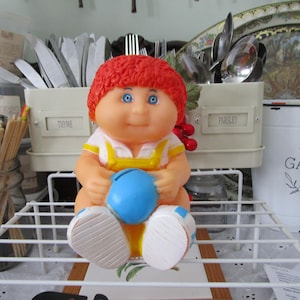 May include: A red-haired Cabbage Patch Kid doll wearing a yellow and blue outfit, holding a blue ball. The doll is sitting on a white wire rack.
