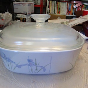 May include: A white casserole dish with a clear glass lid. The dish features a floral design with blue and purple irises. The lid has a clear handle. The dish is on a white surface.