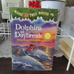May include: A Magic Tree House book titled "Dolphins at Daybreak" by Mary Pope Osborne. The cover features a colorful illustration of two children riding dolphins in the ocean at sunset.
