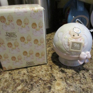 May include: A white porcelain Precious Moments ornament with a baby angel holding a chalkboard with the number 96 on it. The ornament is decorated with a pink bow and the year 1996 is printed on the base.