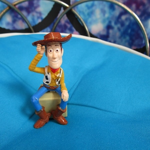 Pode incluir: Uma pequena e colorida figura do personagem Woody do filme de anima&ccedil;&atilde;o Toy Story. Woody est&aacute; usando um chap&eacute;u de cowboy marrom, camisa amarela, colete, cal&ccedil;as azuis e botas marrons. Ele est&aacute; sentado em um pequeno bloco bege, com a m&atilde;o direita levantada para o chap&eacute;u.