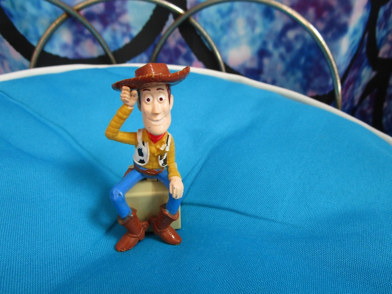 Pode incluir: Uma pequena e colorida figura do personagem Woody do filme de anima&ccedil;&atilde;o Toy Story. Woody est&aacute; usando um chap&eacute;u de cowboy marrom, camisa amarela, colete, cal&ccedil;as azuis e botas marrons. Ele est&aacute; sentado em um pequeno bloco bege, com a m&atilde;o direita levantada para o chap&eacute;u.
