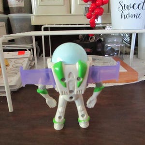 K&ouml;nnte beinhalten: Eine wei&szlig;e und gr&uuml;ne Plastik-Spielzeugfigur von Buzz Lightyear aus der Toy Story-Filmreihe. Die Figur hat einen hellblauen Helm, lila Fl&uuml;gel und ein gr&uuml;nes Brustst&uuml;ck. Die Figur steht auf einer braunen Oberfl&auml;che.