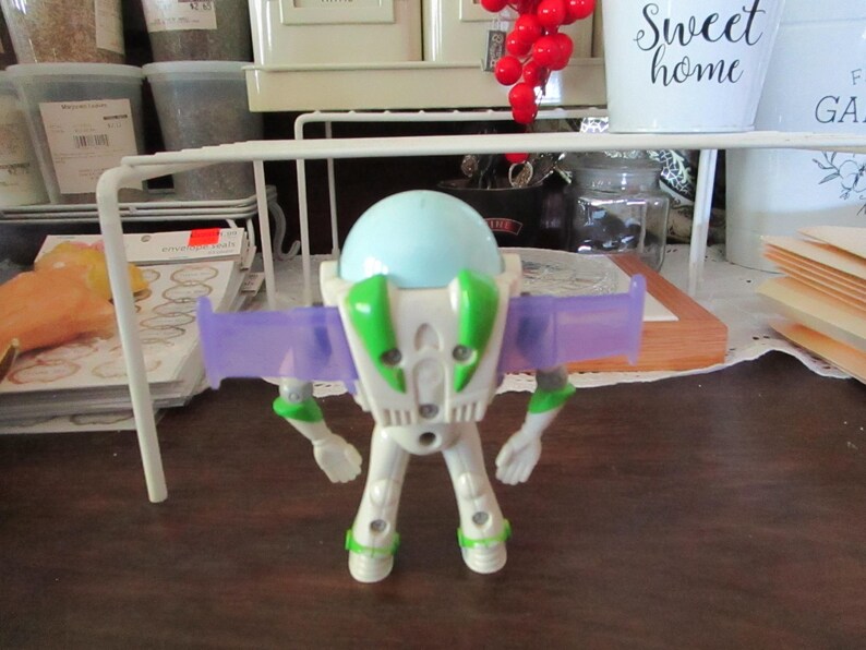 K&ouml;nnte beinhalten: Eine wei&szlig;e und gr&uuml;ne Plastik-Spielzeugfigur von Buzz Lightyear aus der Toy Story-Filmreihe. Die Figur hat einen hellblauen Helm, lila Fl&uuml;gel und ein gr&uuml;nes Brustst&uuml;ck. Die Figur steht auf einer braunen Oberfl&auml;che.