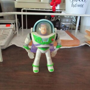 K&ouml;nnte beinhalten: Eine Plastik-Actionfigur von Buzz Lightyear aus der Toy Story-Reihe. Die Figur tr&auml;gt einen wei&szlig;en und gr&uuml;nen Raumanzug mit einem lila Umhang. Die Figur hat einen blauen Helm mit einem klaren Visier und einen roten Knopf auf der Brust.