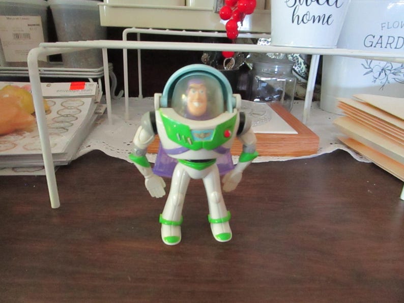 K&ouml;nnte beinhalten: Eine Plastik-Actionfigur von Buzz Lightyear aus der Toy Story-Reihe. Die Figur tr&auml;gt einen wei&szlig;en und gr&uuml;nen Raumanzug mit einem lila Umhang. Die Figur hat einen blauen Helm mit einem klaren Visier und einen roten Knopf auf der Brust.