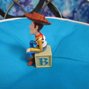 Pode incluir: Uma pequena figura de um personagem de desenho animado de cowboy usando um chap&eacute;u marrom, sentado em um bloco bege com a letra "B". A figura tem uma camisa branca, mangas amarelas, cal&ccedil;as azuis e botas marrons. Fundo azul claro.