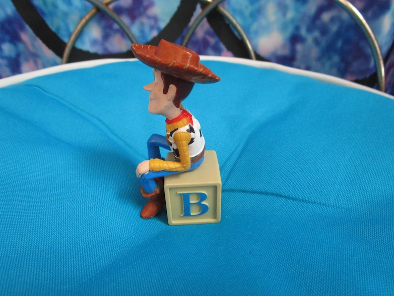 Pode incluir: Uma pequena figura de um personagem de desenho animado de cowboy usando um chap&eacute;u marrom, sentado em um bloco bege com a letra "B". A figura tem uma camisa branca, mangas amarelas, cal&ccedil;as azuis e botas marrons. Fundo azul claro.