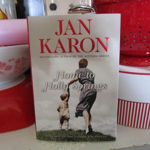 May include: A book titled "Home to Holly Springs" by Jan Karon. The cover features a woman and child walking on grass. The title and author's name are in red. The book is part of the Father Tim Novels series.